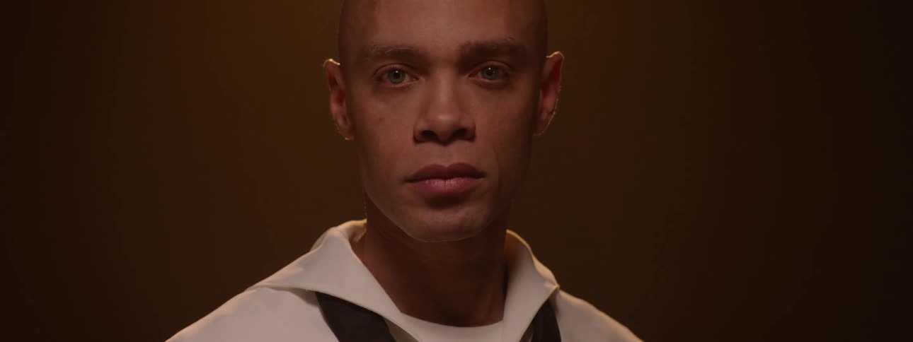 A bald person in a white collared shirt with dark straps looks down and then slowly lifts their head to gaze directly at the camera with striking blue eyes before looking down again all within a dimly lit amber toned setting