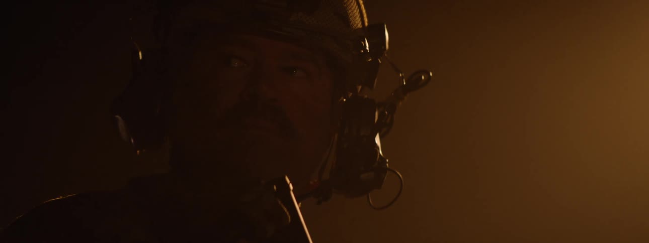 A soldier with a mustache wearing a helmet and headphones turns his head from the side to look directly at the viewer in a dark dusty environment