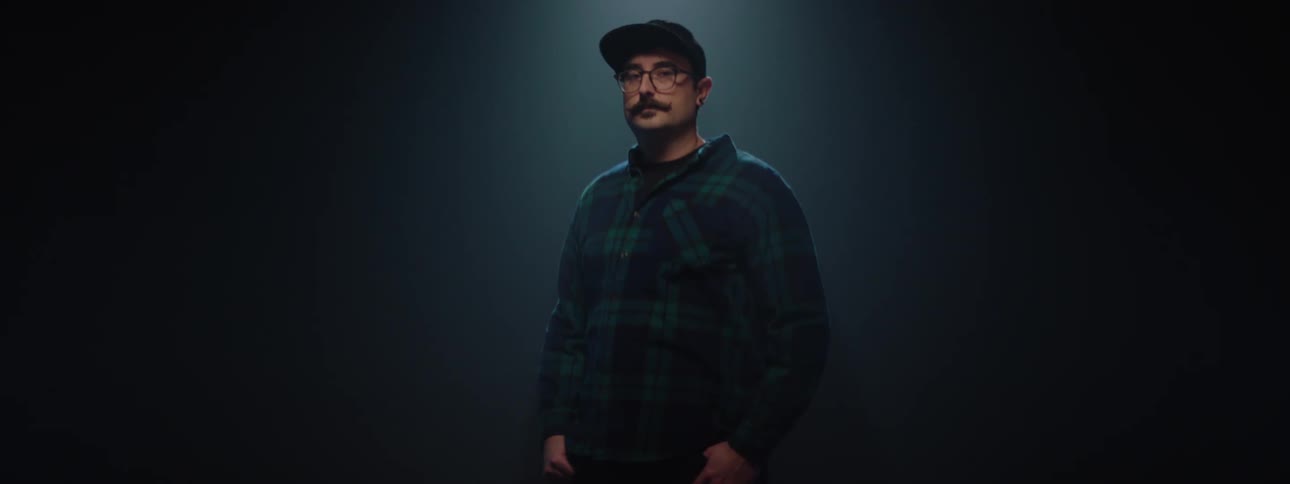 A man wearing a hat long sleeves and glasses standing on a stage