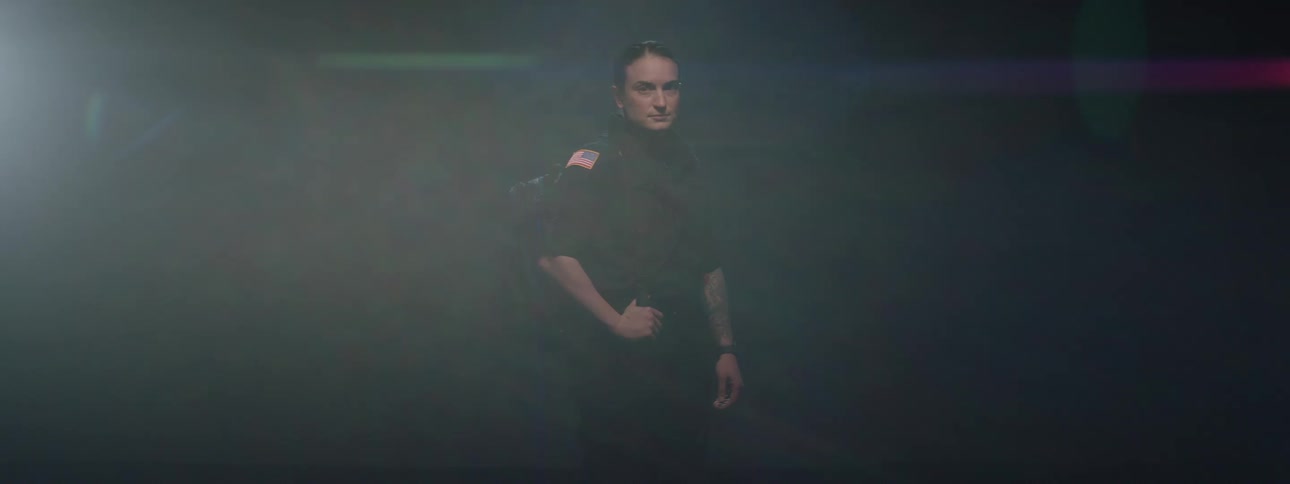 A young woman stands on a dark stage gradually illuminated by light with both hands resting on her waist wearing a police uniform