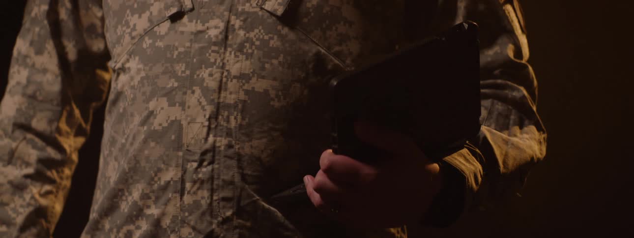 A person wearing military clothing and is holding a small laptop in his hand in an indoor environment with a yellow light in the background
