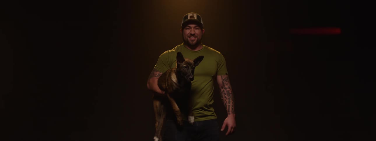 A man wearing a cap and a t shirt with visible arm tattoos turns to face the camera while holding a dog before turning away again all against a dark background with hints of ambient light