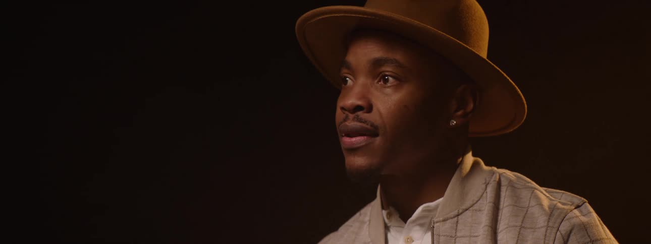The video features a person in silhouette wearing a hat and jacket in a dimly lit setting with a goldenorange light illuminating them from the side as the video progresses they slowly lift their head revealing their face and a gentle smile