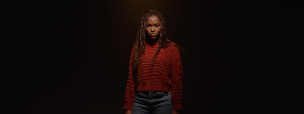 A dark shot of a person with long braids looking directly into the camera as light slowly illuminates their face before the light fades and the person turns to the side