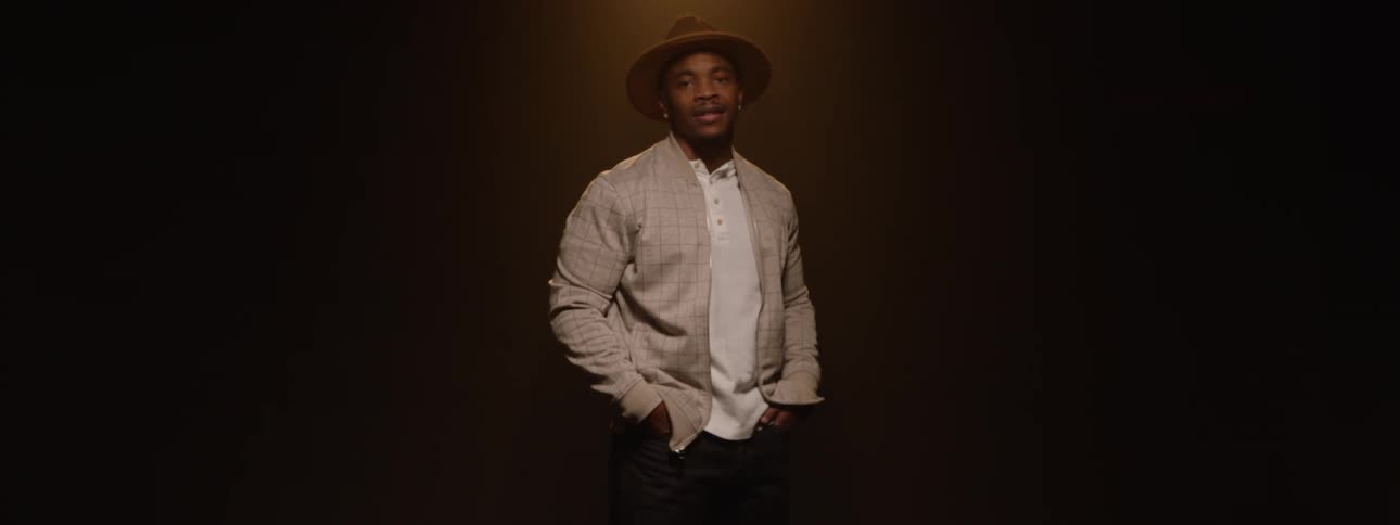 A man wearing formal clothes and a fedora hat and in a dark room with a spotlight and he spinning it around him