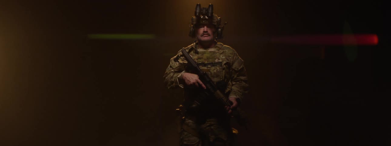 Older soldier wears battle gear and carries a rifle while rotating in place in a hazy area lit by a spotlight