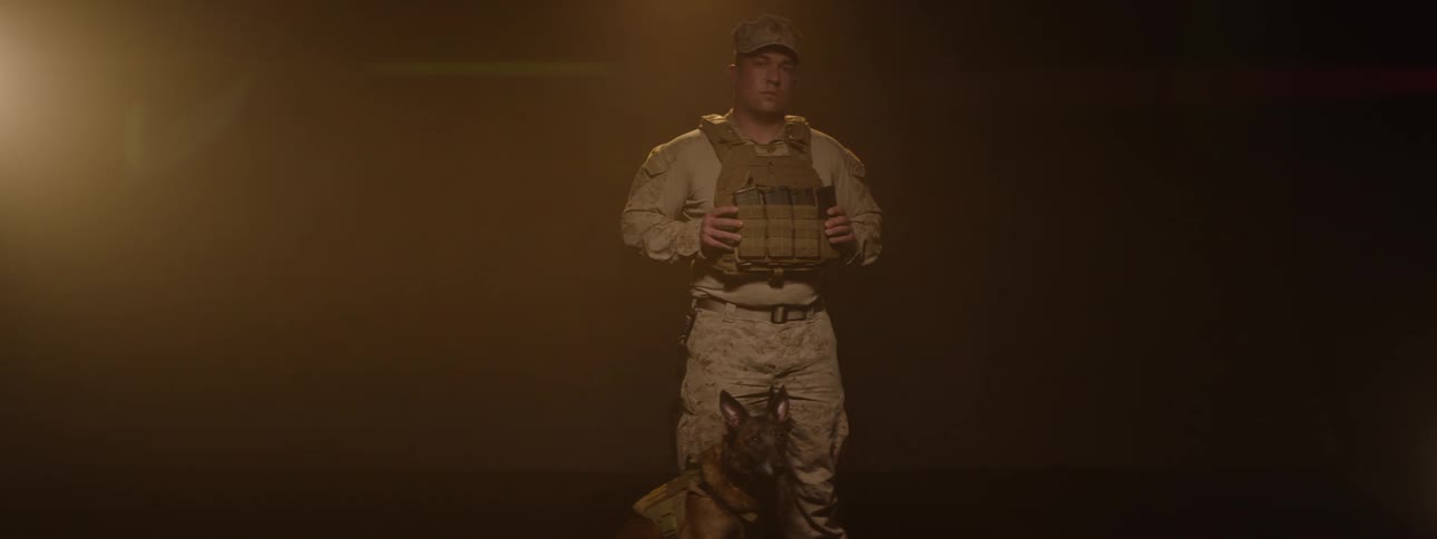 The video features a soldier in full camouflage uniform and tactical vest standing in a dimly lit smoky environment suggesting a dramatic or intense setting a military working dog is visible sitting at his feet completing the powerful image