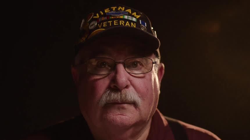 Close up portrait of a middle aged man wearing a black veteran cap with glasses in a dark room