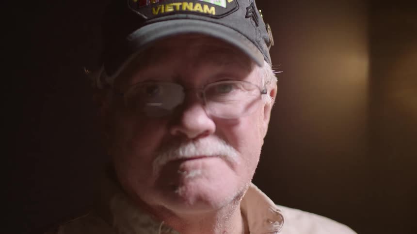 The video shows an older man with white hair and a white mustache wearing glasses and a dark baseball cap that says vietnam he looks directly at the camera with a serious and contemplative expression his face illuminated by a soft