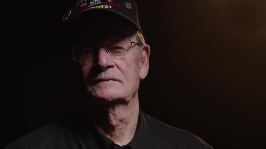 A man looks up at the camera while wearing a veterans hat