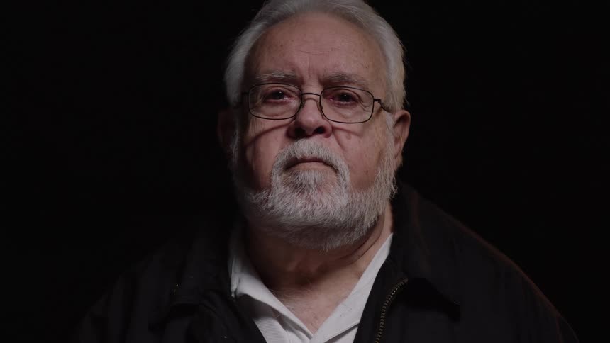A military officer with a white beard wearing glasses looking straight ahead indoors