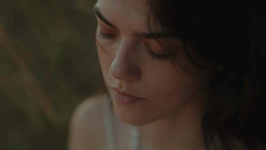 Young dark haired female with blue eyes sitting in tall grass she has a longful look on her face while looking around