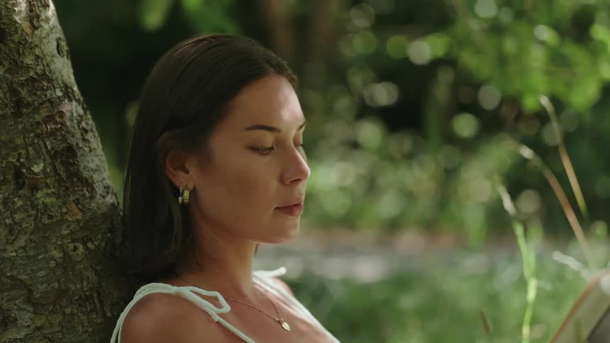 Woman reads a book leaning back against a tree