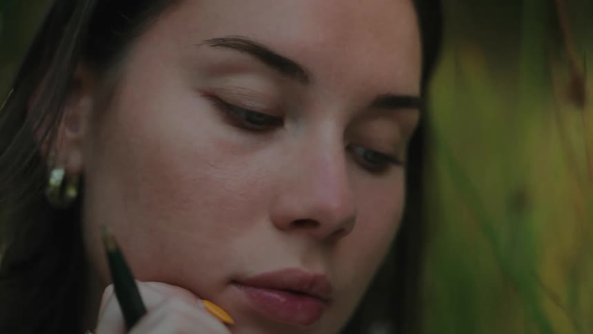 A young woman wearing round earrings sitting beside a wall with a pen on her arm lost in thought