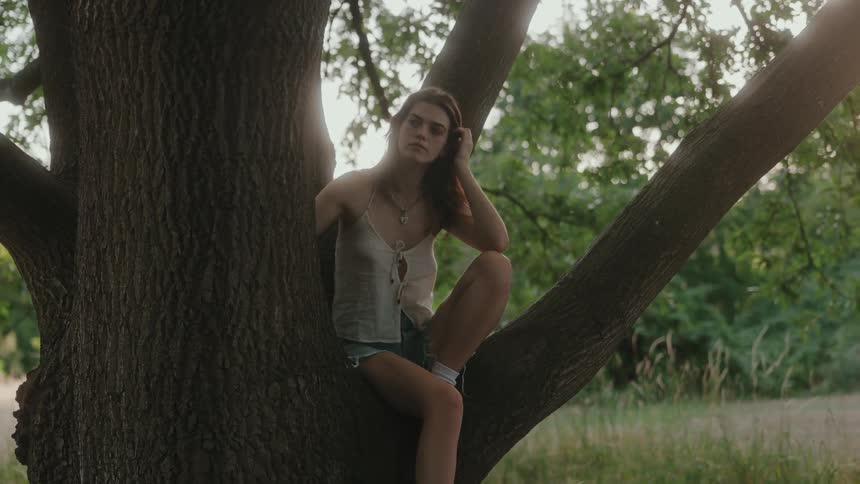 A young woman sitting on a tree trunk in the woods at midday rubbing her long dark hair