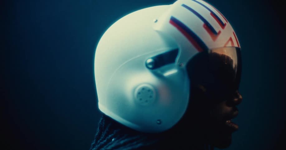 A black man with long hair wears a futuristic white helmet with red and blue stripes and a dark visor shown in dramatic side profile lighting