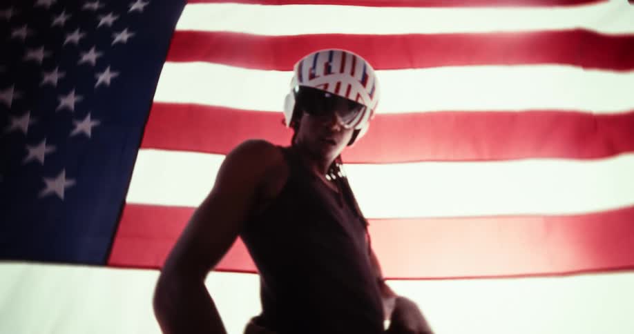 The video features a person wearing a white helmet with red and blue stripes and sunglasses standing in front of an american flag they are seen gesturing with their hands at one point pointing directly at the camera
