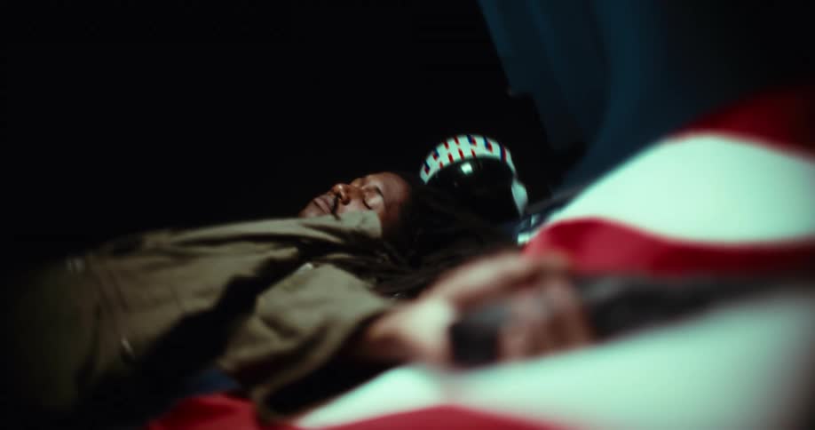 The video shows an individual with long dark hair lying down possibly in a state of rest or mourning a blurred american flag is partially visible in the foreground suggesting a patriotic or commemorative context
