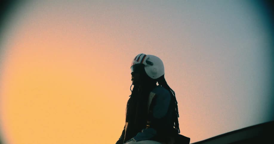 A black person with long braids wearing a white helmet with red and blue stripes sits on a rooftop at sunset silhouetted against a warm orange sky