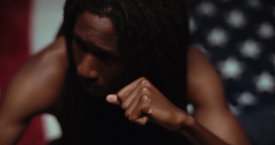 The video shows a closeup of a person with dark skin and dreadlocks wearing a dark tank top in front of what appears to be an american flag they initially look towards the camera then lower their head and bring their hand to their face