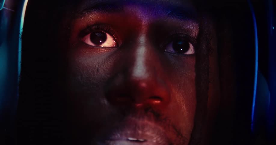 A black person with a helmet on stares upward with wide focused eyes and talking is bathed in colorful light