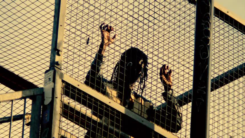 A black person with long braids grips a wire fence backlit dramatically by daylight