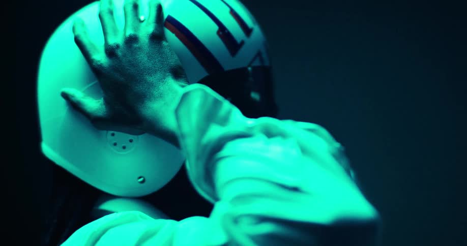 A black person with long braids adjusts their helmet under vivid teal lighting