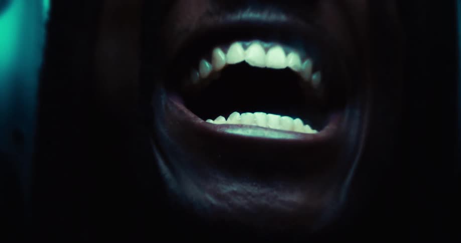 The video shows an extreme closeup of a persons mouth wide open revealing their teeth the lighting is dark with a blue or greenish hue illuminating the teeth and the inside of the mouth