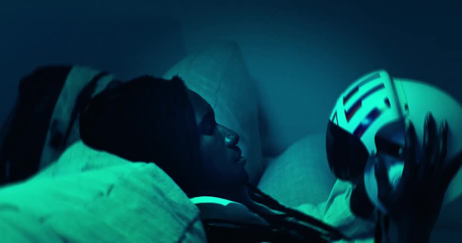 The video shows a person lying down looking towards the right side of the frame bathed in a strong blue light they are holding a white helmet with red and blue stripes on the side bringing it into the frame from the right