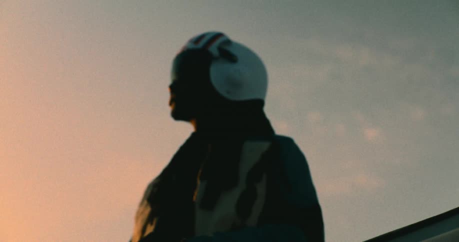 A silhouette of a black person in a helmet stands against a soft sunset sky