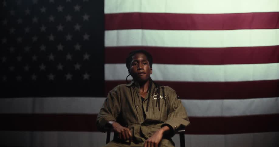 A spotlight comes on as soon as a man sits in a chair in front of an american flag