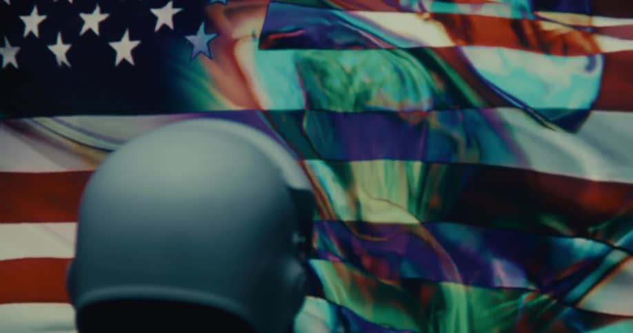 A man wearing a helmet stares at a large screen displaying the american flag