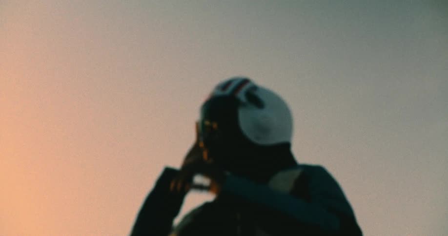 A motorcycle rider wearing a leather jacket and a helmet standing under the setting sun