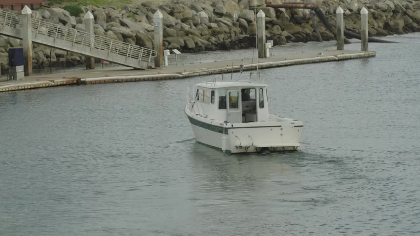A small white motorboat approaches a dock in a calm harbor with rocky breakwaters and a ramped pier in the background
