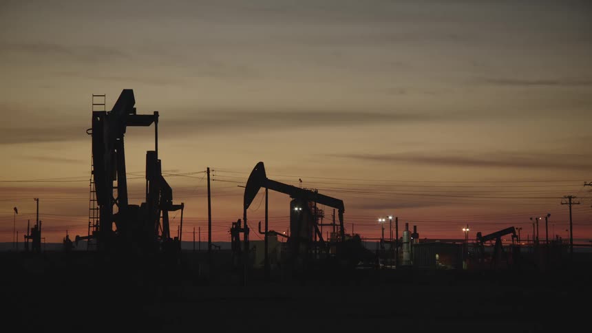 Silhouetted oil field at dusk with several large oil pumpjacks standing tall against a warm fading sunset sky