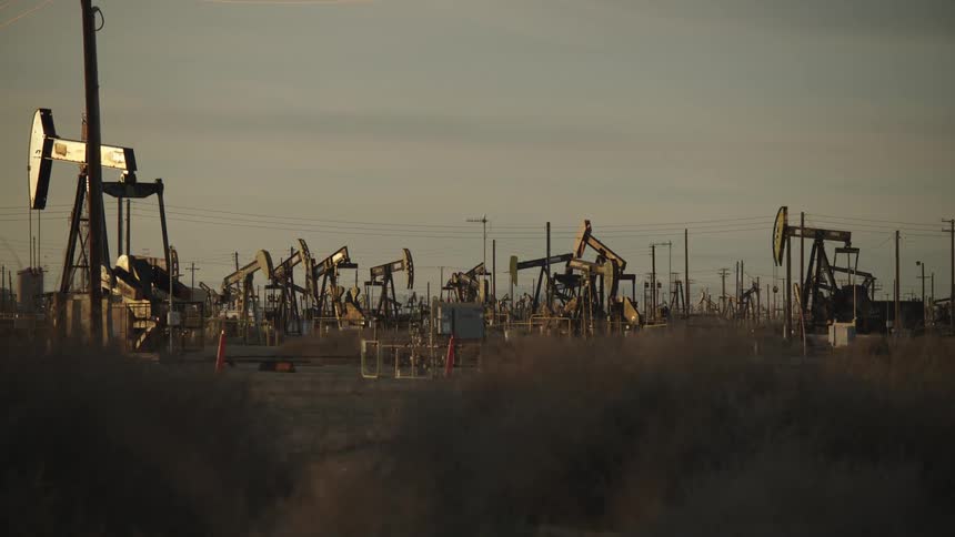 Rows of oil pump jacks stretch across a dusty barren field under a dim overcast sky creating a stark and industrial landscape