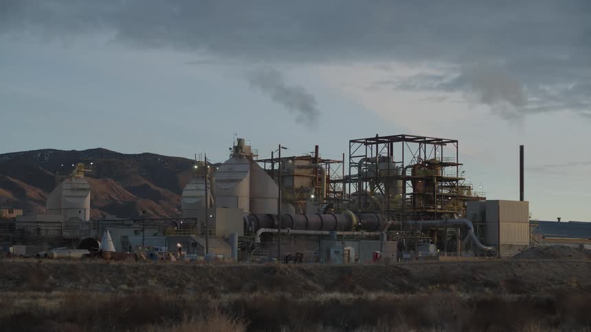 An industrial plant sits at the base of arid rugged hills releasing smoke into a cloudy sky