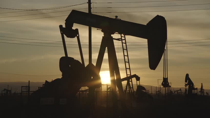 An oil pump jack is silhouetted against a golden sunset with power lines and additional rigs in the background evoking a gritty vintageindustrial mood