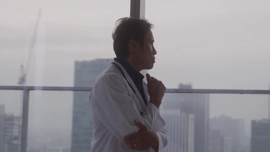 Male medical doctor thinks while walking close to windows showing a foggy city during the daytime