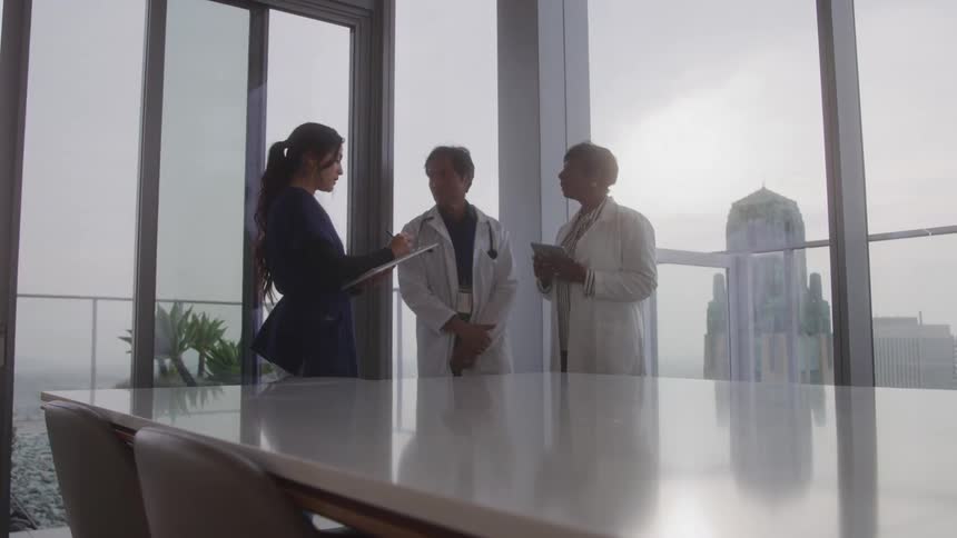Two people in lab coats talking to a woman taking notes in a notebook in an office room