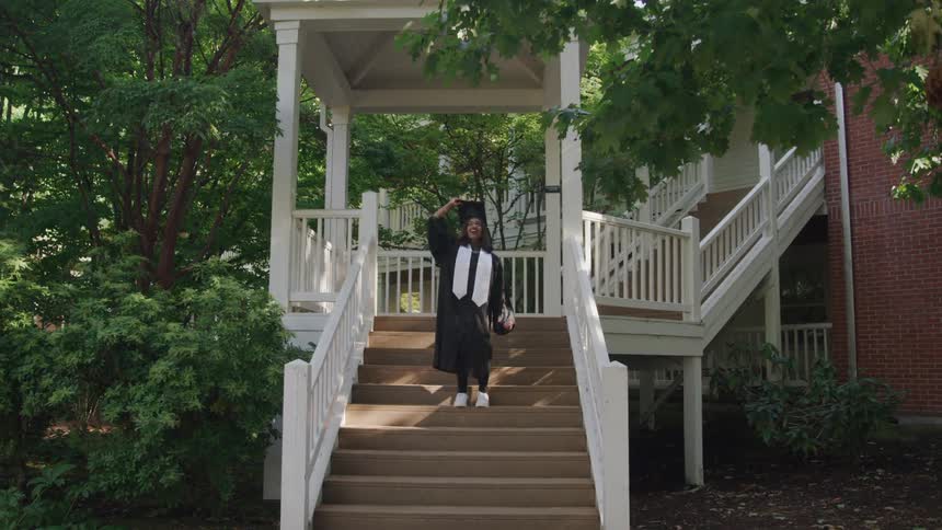 The video shows a graduate in a black cap and gown standing on a wooden staircase with white railings surrounded by lush green trees and a brick building in the background she joyfully throws her graduation cap into the air celebrating her achievement