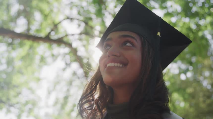 The video features a female graduate in a black cap and gown standing outdoors with a bright joyful expression she is looking upwards and a strong natural light source from above creates a beautiful lens flare enhancing the celebratory mood