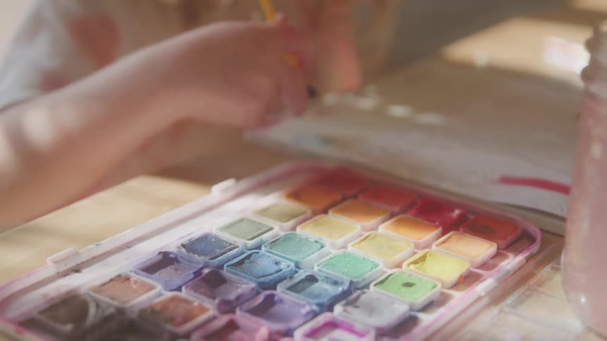 The video shows an extreme closeup of a persons hands as they meticulously mix watercolors in a palette revealing a wide array of vibrant colors the focus then shifts as they bring the brush to a sheet of paper to begin painting