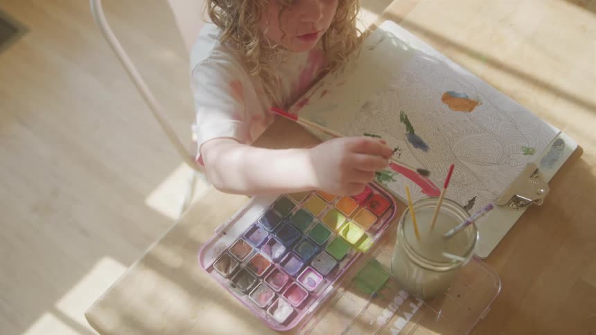 Highangle shot of a young child with curly blonde hair seated at a wooden table and painting with watercolor paint