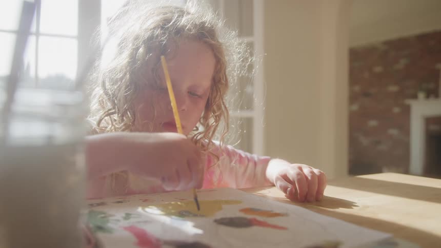A young girl with curly hair paints at a sunlit table surrounded by soft overexposure and haze that gently blur the scenes details