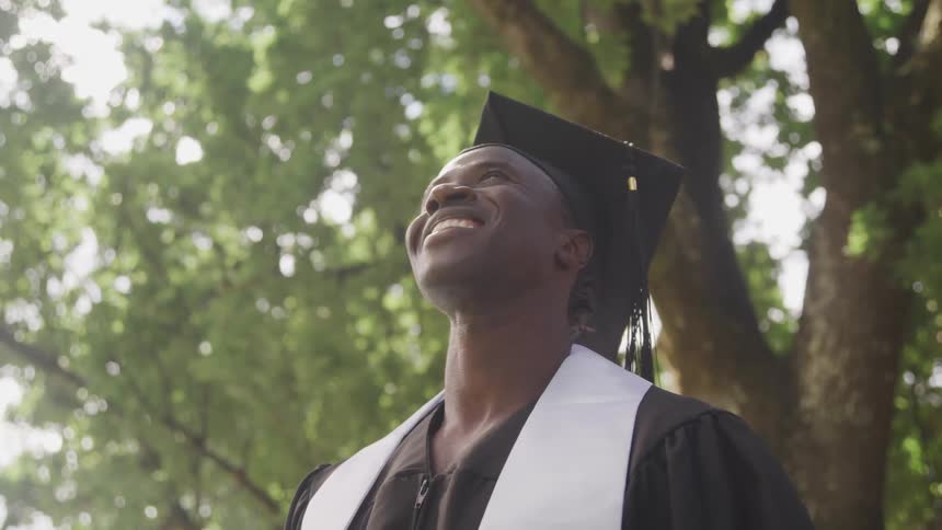 Young man wearing a black graduation cap and gown with a white stole looking upwards with a broad smile