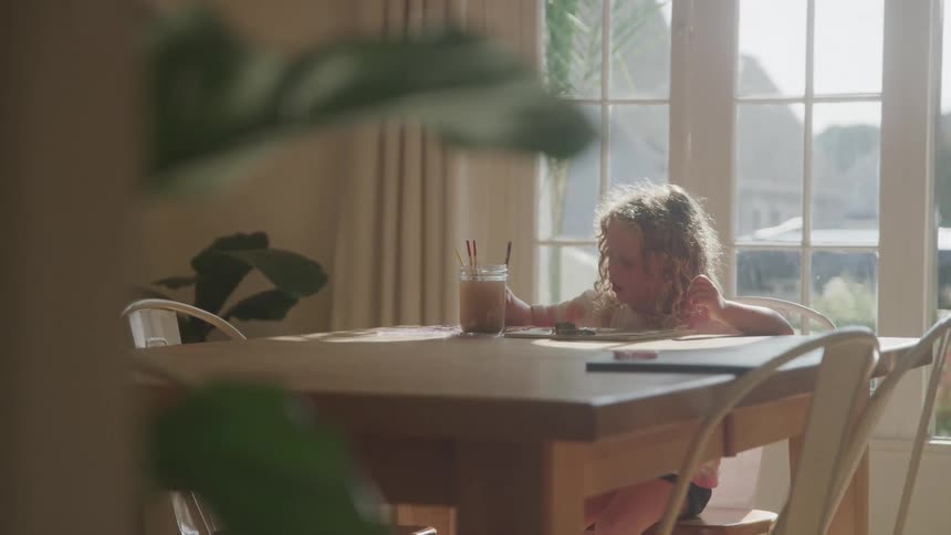 The video features a young child with curly lightcolored hair sitting at a wooden table in a brightly lit room they are focused on an activity holding a long spoon and stirring a dark liquid in a glass