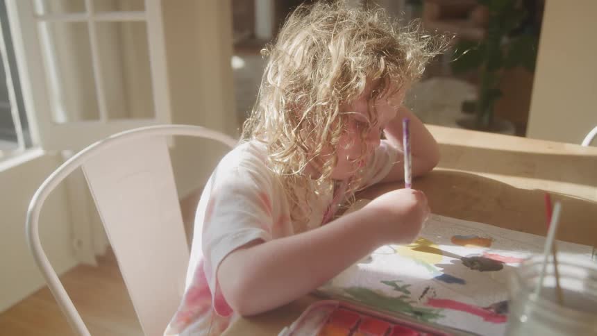 Young child with curly blonde hair sitting at a wooden table engaged in painting with watercolors