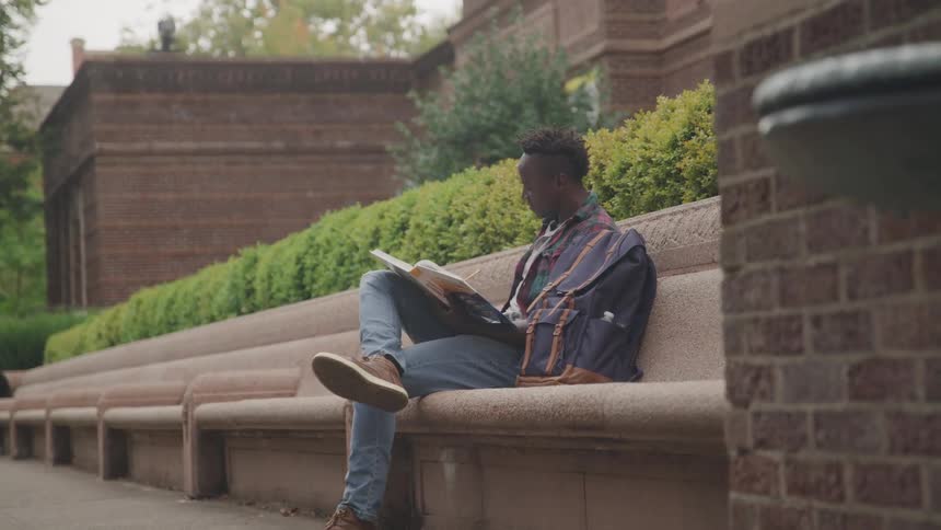 Young black african american man sitting on a long stone bench outdoors studying