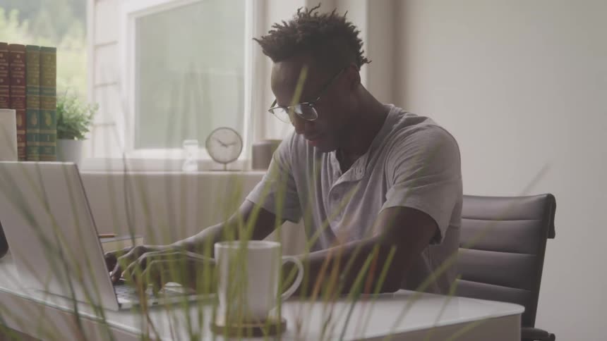 Young man with dark styled hair and glasses seated at a desk and actively working on a laptop